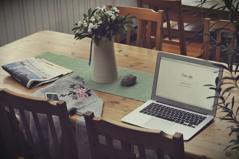 An open laptop showing the Google homepage beside flowers and a newspaper