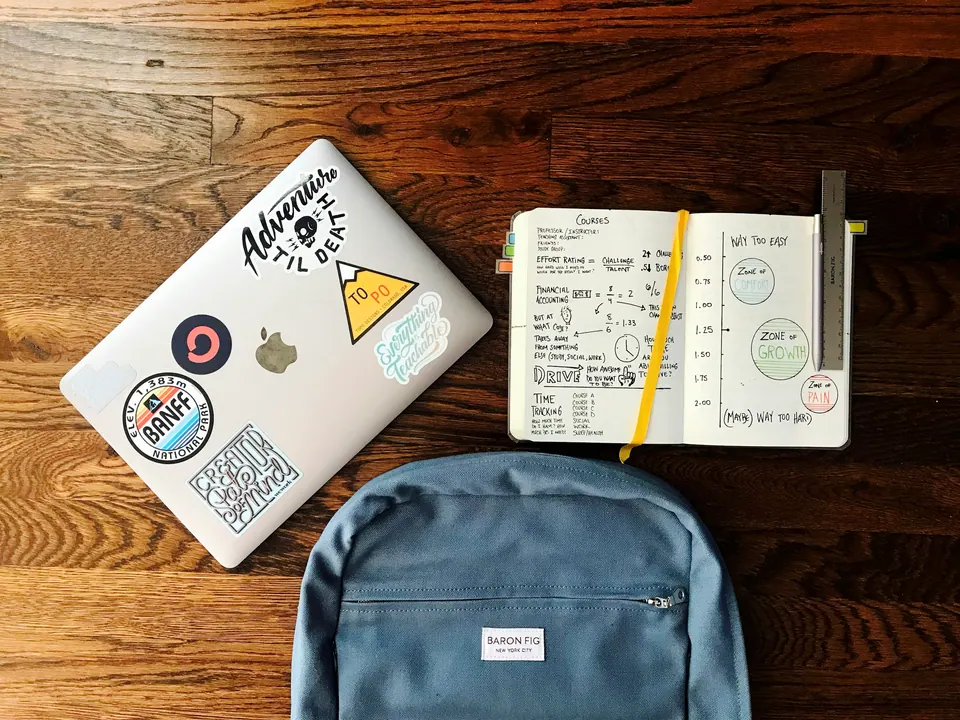 A closed laptop, an open planner and a blue backpack on a wooden surface