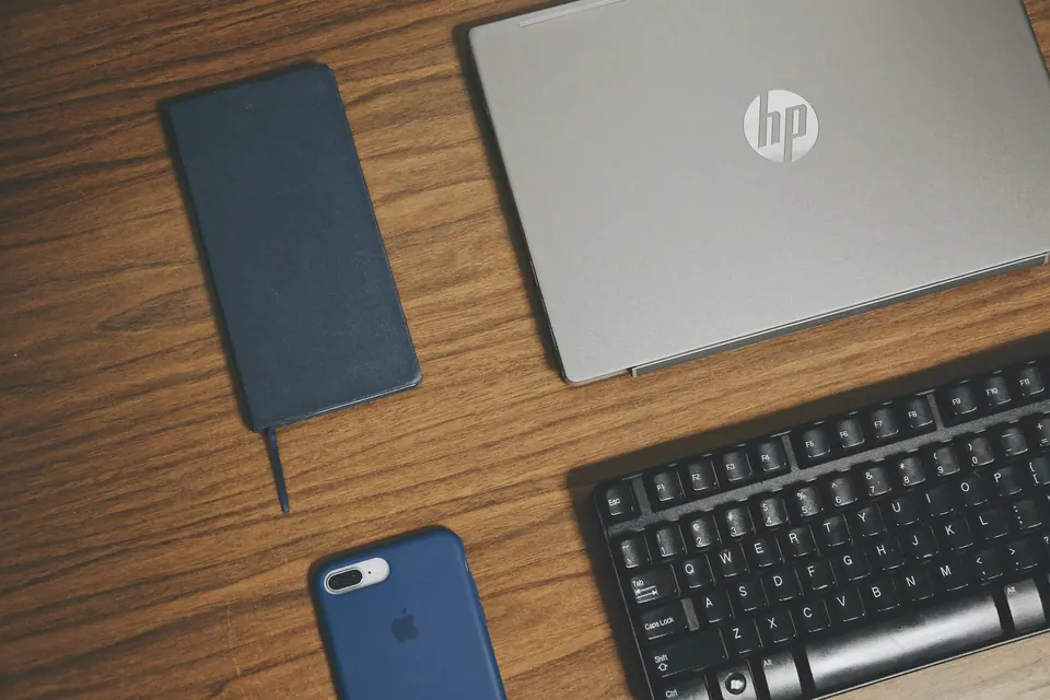 A laptop, keyboard, phone and notebook arranged on a wooden surface