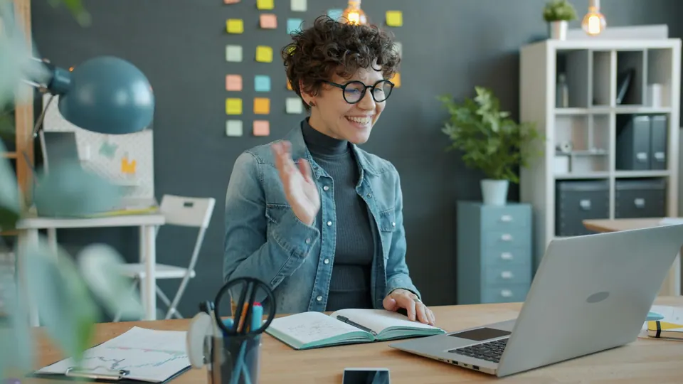 A woman waving towards her laptop during a video call at a desk